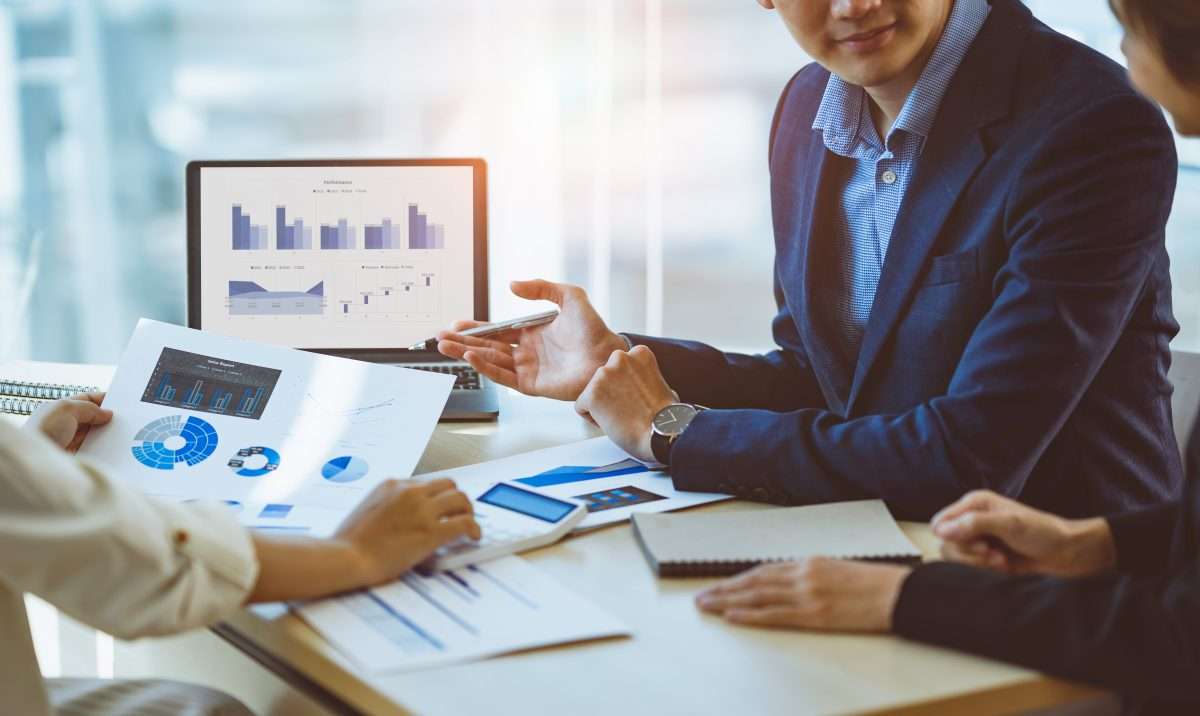 Business professionals reviewing financial charts and performance graphs on a laptop and printed reports during a strategy meeting, illustrating business valuation analysis amid economic uncertainty.